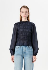 Navy blue smocked blouse with long sleeves, textured fabric, and a ruffled neckline, paired with light blue high-waisted jeans.