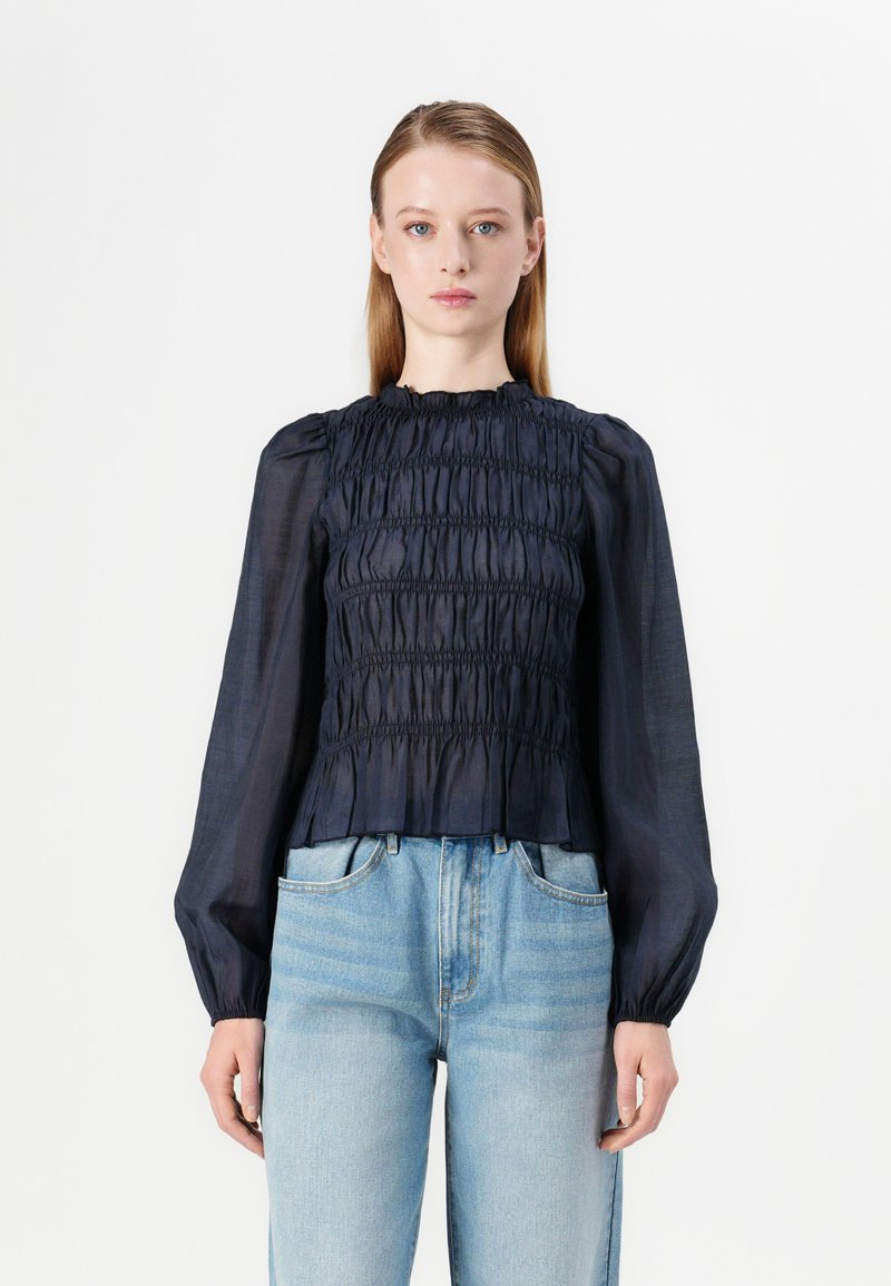 Navy blue smocked blouse with long sleeves, textured fabric, and a ruffled neckline, paired with light blue high-waisted jeans.