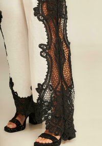 Witte wijde broek met ingewikkeld zwart kant langs de zijkanten, voorzien van een gedurfde bloemenprint en textuuraccenten. Zwarte hoge hakken sandalen.