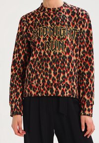 Chemise à manches longues imprimé léopard avec "MIDNIGHT RUN" en lettres jaunes et noires audacieuses, portée avec un pantalon noir.