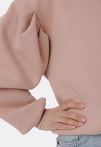 Sudadera en color rosa claro con mangas abullonadas, puños acanalados y textura suave. Mano resting sobre la cadera, combinada con jeans de denim de lavado claro.