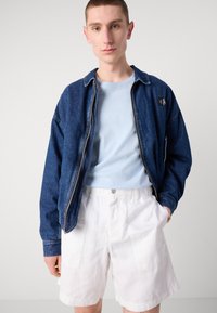 Denim-tyylinen vetoketjullinen takki sinisen T-paidan ja valkoisten shortsien kanssa. Takissa on logo-patch ja rento leikkaus, sekä näkyvät ompelukohdat.
