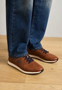 Baskets en cuir marron avec des détails perforés, semelle en caoutchouc blanche et lacets navy, associées à un jean en denim bleu. Fond en bois.