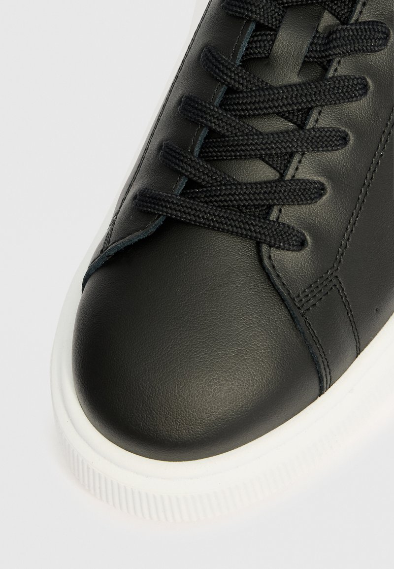 Baskets en cuir noir avec une texture lisse, présentant un bout rond, des lacets noirs et une semelle en caoutchouc blanche avec des rainures verticales.