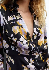 Blouse à motifs floraux sur fond noir, avec des motifs violets et dorés. Elle possède un col et un décolleté en V profond, fabriquée en tissu lisse.