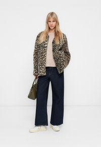 Νεαρή γυναίκα με μακριά ξανθά μαλλιά, φορώντας ένα παλτό με λεοπάρ σχέδιο, ανοιχτό μπεζ πουλόβερ, φαρδιά σκούρα τζιν, λευκά αθλητικά παπούτσια και κρατώντας μια ελιά τσάντα.