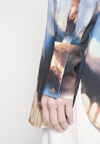 Button-up shirt featuring a tie-dye pattern in blue, brown, and cream tones. It has a structured collar and brown buttons on the cuff.