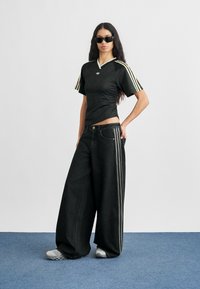 Musta urheilullinen toppi, jossa valkoiset ja kullanväriset raidat hihoissa, yhdistettynä löysälahkeisiin mustiin farkkuihin, joissa on sivuraidat ja teksturoitu pinta.