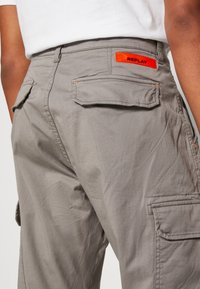 Grijze cargobroek met meerdere zakken, oranje accenten en een "REPLAY" logo-patch aan de achterkant. Gemaakt van gestructureerde katoenen stof.