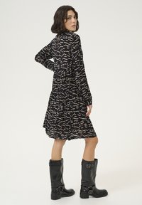Robe noire avec un motif abstrait beige, manches longues, design à volants, et un dos avec détail en trou de serrure, associée à des bottes noires jusqu'aux genoux.