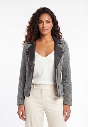 Femme aux cheveux bouclés portant une veste gris chevrons avec des revers noirs, un haut blanc à col en V, un pantalon beige et des boucles d’oreilles créoles dorées.