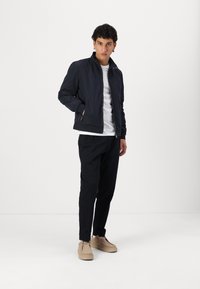 Marine bomberjack met een staande kraag, ritssluiting en zijzakken, gedragen over een wit shirt, gecombineerd met zwarte broek en beige schoenen.
