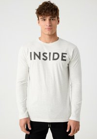 Camiseta de manga larga gris claro con cuello redondo, que presenta un texto negro en negrita "INSIDE" en la parte frontal. Tejido suave con una textura lisa.