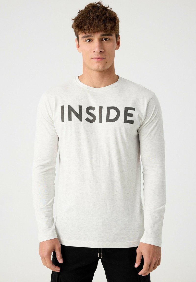 Camiseta de manga larga gris claro con cuello redondo, que presenta un texto negro en negrita "INSIDE" en la parte frontal. Tejido suave con una textura lisa.