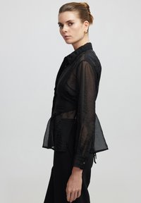 Camicia nera trasparente con maniche lunghe, chiusura a bottoni frontale e dettagli arricciati in vita. Il tessuto ha una texture stropicciata.