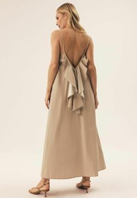 Beżowa maxi sukienka z niskim tyłem i warstwowymi falbanami, wykonana z lekkiego materiału. Posiada cienkie ramiączka i miękką fakturę.