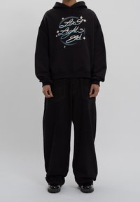 Sudadera negra con capucha y un gráfico frontal, con texto en azul y blanco. Combina con pantalones negros holgados y calzado negro brillante.