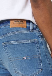 Blå denimjeans med en läderetikett som säger "TOMMY JEANS" och en liten logotyp på bakfickan. Klassisk femficksdesign.