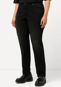 Jeans de denim negro con corte recto, con un ligero desgaste. Combinados con zapatos de plataforma negros, llevados por una persona frente a un fondo neutro.
