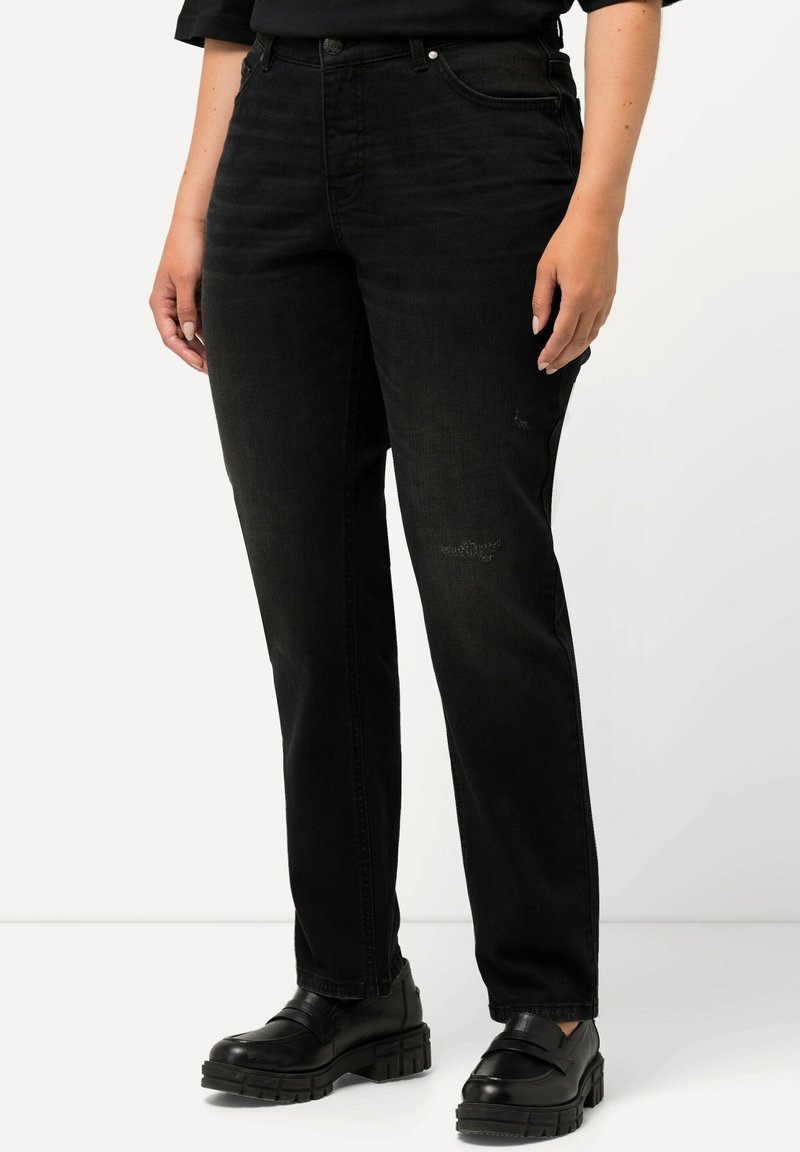 Jeans de denim negro con corte recto, con un ligero desgaste. Combinados con zapatos de plataforma negros, llevados por una persona frente a un fondo neutro.