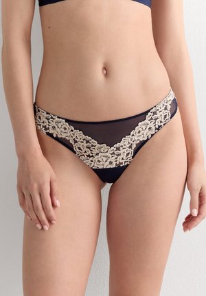 Žena v černých průsvitných kalhotkách s béžovou květinovou krajkou, stojící před jednobarevnou světlou zdí.