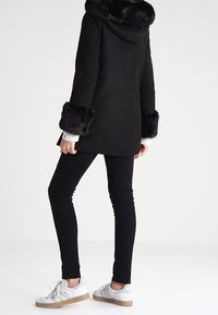 Černý kabát s kapucí z umělé kožešiny a lemováním na rukávech, s přiléhavým střihem, nošený přes černé skinny džíny a bílé tenisky.