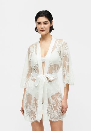 Noor naine lühikeste tumedate juustega, kandes läbipaistvat valget pitsist hommikumantlit, mis on talje juures kinni seotud ja mille all on valged pesud, seistes valge tausta ees.