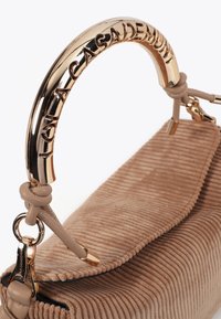 Una borsa beige strutturata con un manico dorato lucido che presenta la frase incisa "Hola Casademunt." La borsa ha una superficie morbida e rigata.