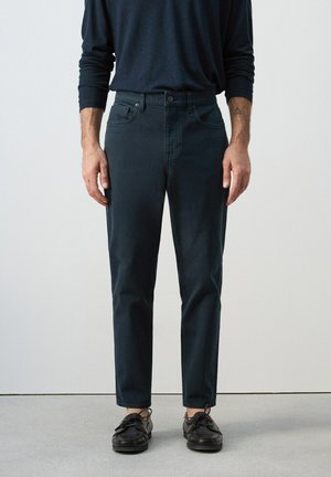 Uomo che indossa pantaloni scuri aderenti, scarpe da barca nere e una camicia a maniche lunghe scura, in piedi contro uno sfondo chiaro e uniforme.