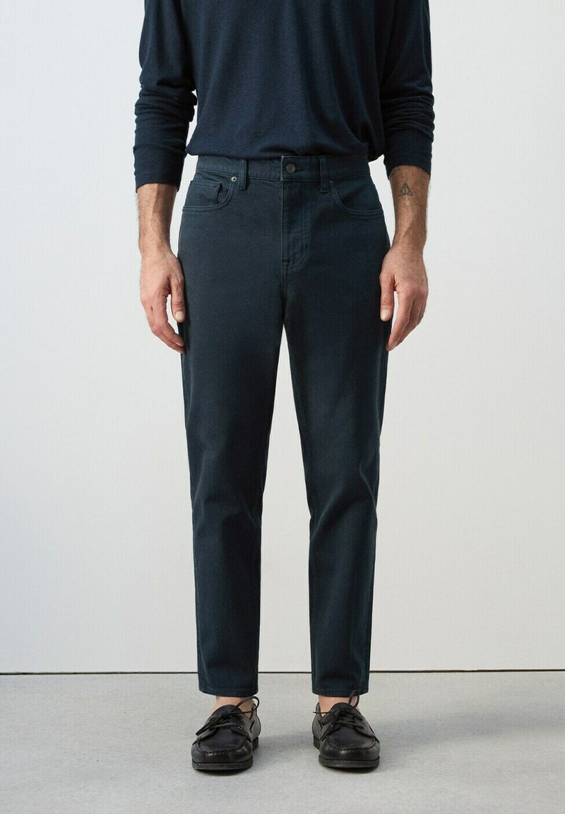 Muž oblečený v tmavých úzkych nohaviciach, čiernych lodných topánkach a tmavom tričku s dlhým rukávom stojaci pred jednoduchým svetlým pozadím.