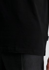 Schwarzes T-Shirt mit Saum und kleinem Seitenschild mit der Aufschrift "pierre cardin" über dunkelgrauen strukturierten Hosen, mit einem Teil einer Hand auf der rechten Seite sichtbar.