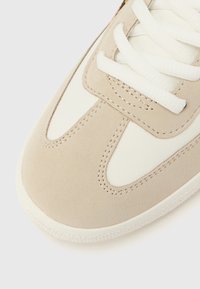 Μπεζ και λευκό αθλητικό παπούτσι με υφή σουέντ στη μύτη, λεπτομερείς ραφές και λευκά κορδόνια σε απλό φωτεινό φόντο.