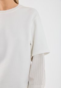 Witte sweatshirt met korte, losse mouwen, gelaagd over een licht gestreepte lange mouw. Zachte textuur, casual ontwerp en minimalistische stijl.