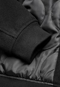 Black fleece and quilted fabric jacket featuring ribbed cuffs and visible stitching patterns on the exterior. Soft texture with a matte finish.