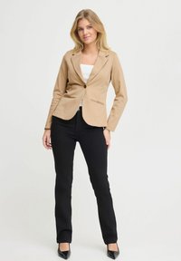 Blazer beige su misura con un unico bottone marrone, due tasche frontali e una vestibilità slim, abbinato a pantaloni neri e tacchi neri eleganti.