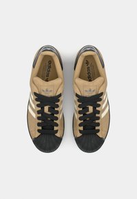 Tan suede sneakers with black rubber toe cap, three white stripes, and black laces. Features a padded collar and textured sole.