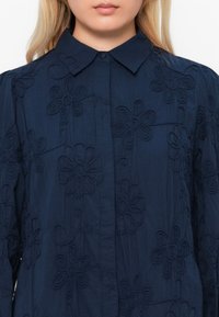 Navy blue shirt with a button-down collar, featuring embroidered floral patterns and a textured fabric. Displaying a relaxed fit design.
