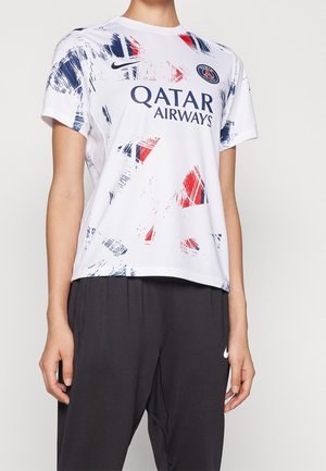 Personne portant un maillot de football blanc du Paris Saint-Germain avec des motifs rouges et bleus imitant des coups de pinceau et un pantalon noir Nike.