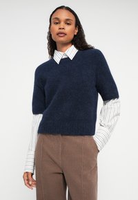 Nő, aki tengerészkék rövid ujjú pulóvert visel fehér csíkos ing felett és barna kordbársony nadrágot, egy sima fehér háttér előtt áll.