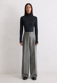 Eine Frau mit langen, glatten Haaren steht vor einer schlichten Wand und trägt einen schwarzen Rollkragenpullover, graue, weite Hosen und schwarze Schuhe.