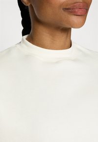 Valge kõrge kaelusega sweatshirt, pehme tekstuuriga ja minimaalse õmbluste detailiga. Suurendatud vaade keskendub kaeluse ja kangakvaliteedi detailidele.