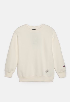 Sweatshirt couleur crème en tissu doux, avec un col rond, des poignets côtelés et une petite patch logo sur le bas à gauche.