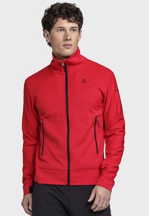 Man wearing a red zip-up jacket with black zipper pockets, standing and looking slightly right on a light background.