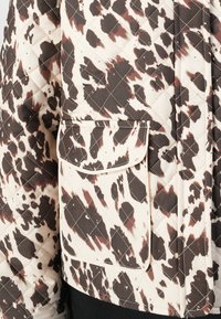 Chaqueta acolchada con una base crema y un estampado abstracto en marrón oscuro y marrón claro. Incluye un bolsillo frontal con solapa.