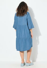 Denimimekko, jossa on kerrostettu muotoilu, kolmanneksen hiha ja pyöreä helma. Vaaleansininen kangas pehmeällä tekstuurilla. Yhdistetty sinisiin puukkoihin.