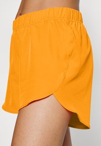 Kirkkaanoranssit shortsit, joissa on joustava vyötärönauha, väljä istuvuus ja kaareva helma. Kangas näyttää sileältä ja kevyeltä.