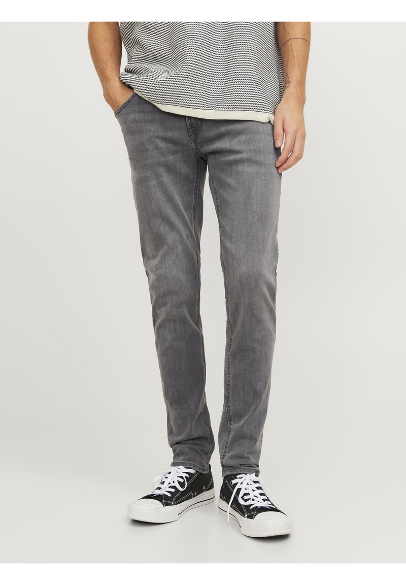 Jeans de mezclilla gris con un corte slim, diseño de cinco bolsillos y un sutil desgaste. Combinados con zapatillas negras con cordones blancos.