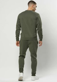 Mann stående med ryggen til, iført olivengrønn genser og matchende joggebukser med hvite joggesko på ensfarget bakgrunn.