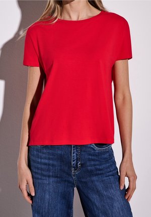 Woman wearing a plain red short-sleeve T-shirt and blue jeans, standing against a gray wall with hands relaxed at sides.