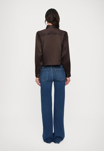 Hnedá saténová košeľa s golierom a dlhými rukávmi, skrátený strih. Kombinovaná s modrými rozšírenými džínsami s vreckami na zadnej strane a hladkou textúrou.